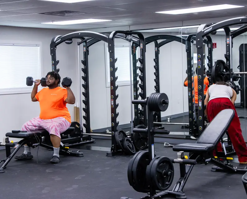 Two people lifting dumbbells beside benches and racks in a fitness room